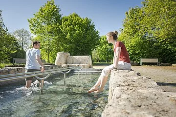 Pärchen beim Wassertreten im Kurpark. Frau schaut zu und Mann tritt Wasser.