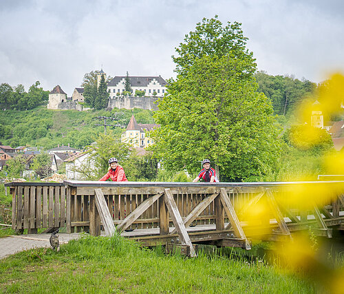 Ein Ehepaar überquert per Rad die Holzbrücke in Möhren. Im Hintergrund sieht man das Schutzengelhaus und das Schloss Möhren