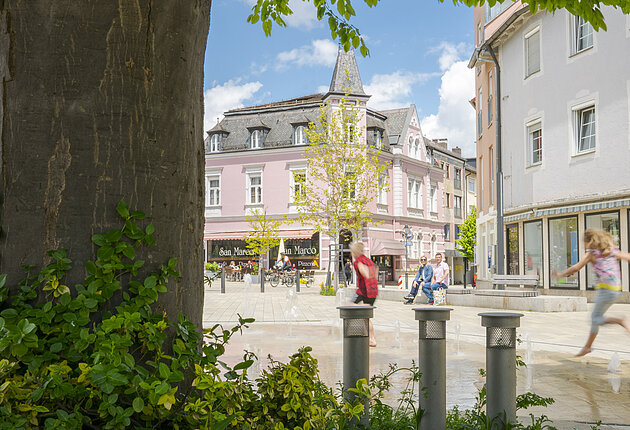 Blick unter den Blättern des Bäumes hin zum Brunnen am Wallmüllerplatz, Kinder springen im Wasser, im Hintergrund sieht man das historische Gebäude von Fam. Fuchs