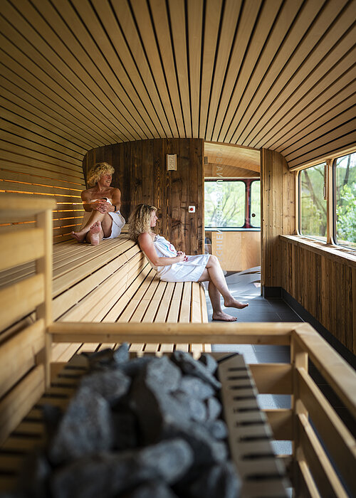 Zwei Frauen sitzen in der Schienenbus-Sauna mit dem Blick ins Freie. Im Vordergrund ist der Saunaofen