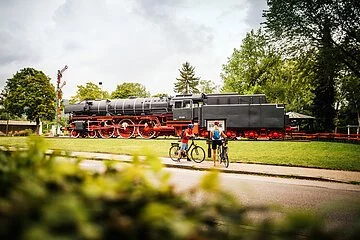 Zwei Radler kommen gerade an der Eisenbahn, die in Treuchtlingen steht, vorbei. Die Bahn ist inmitten einer grünen Wiese zu finden.