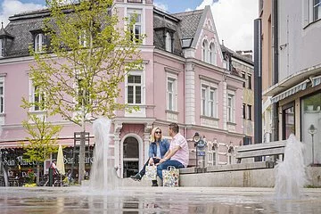 Ehepaar sitzt auf einer Bank am Wallmüllerplatz vor den Wasserspielen. Im Hintergrund gross das historische Gebäude von Familie Fuchs.