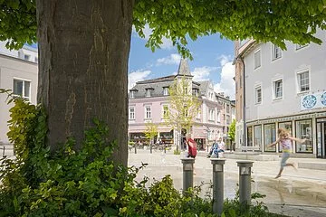 Blick unter den Blättern des Bäumes hin zum Brunnen am Wallmüllerplatz, Kinder springen im Wasser, im Hintergrund sieht man das historische Gebäude von Fam. Fuchs