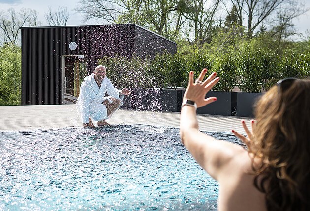 Gast spritzt im Außenbecken der Sauna gegenüber sitzende Frau voll