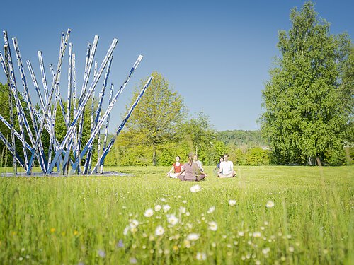 Yoga in der Gruppe im Kurpark. Kunstwerk mit langen Stangen und Bäume sind im Bild Hauptakteur
