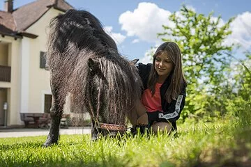Grasendes Pony und kniendes Mädchen. Im Hintergrund ein Bauernhof und ein großer Baum