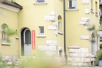 Altes Zollhaus aus Perspektive des gegenüberliegenden Gehsteigs. Dieses Gebäude ist geprägt von weissen Steinornamenten an den Kanten des leicht grünen Gebäudes.