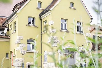 Das alte Zollhaus mit Blick auf die Front des leicht grünen Gebäudes aus Sicht des gegenüberliegenden Gehsteigs. Das Gebäude ist geprägt von weissen Steinornamenten. Im Vordergrund des Bilder sind Pflanzen zu sehen.