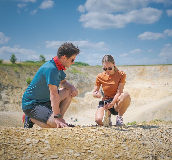Ein Mann im blauen Shirt kniet neben einer Frau, die mit Hammer und Meißel vorsichtig eine Steinplatte im Fossiliensteinbruch Blumenberg teilt.