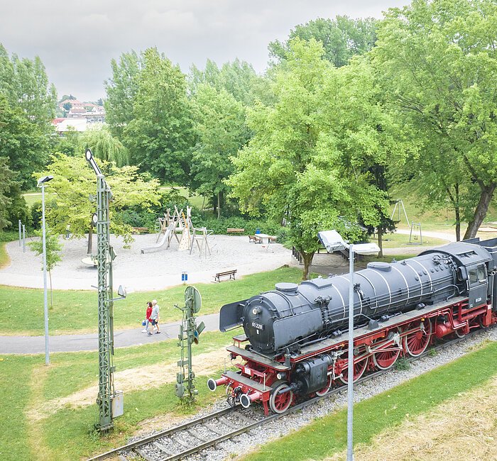 Luftaufnahme der Treuchtlinger Denkmalslok inklusive dahinter liegendem Spielplatz mit Besuchern inmitten von Grün.