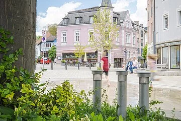 Wasserspiele am Wallmüllerplatz, eingerahmt von Pflanzen im Vordergrund und Baum. Im Hintergrund historisches Gebäude der Fam. Fuchs