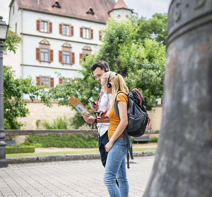 Zwei Touristen laufen auf der Straße zwischen Museum und Stadtschloss Treuchtlingen. Sie lauschen der Stadttour über die Kopfhörer der Audioguides. Im Hintergrund ist das Treuchtlinger Stadtschloss zu sehen.