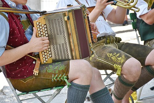 Mann mit Lederhosen sittz auf einem Stuhl und spielt Akkordeon.
