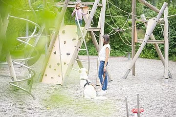 Moderner Spielplatz an der Denkmalslok. Ein Kind klettert auf den aus Holz hergestellten Geräten. Mutter mit Hund stehen davor und beobachten das Kind.