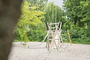 Kleiner, moderner Spielplatz mit Spielelementen aus holz auf feinem Kieselbankett inmitten von grünen Bäumen.