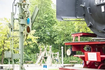 Angeschnittene Aufnahme der Denkmalslok im seitlichen Vordergrund. Im Hintergrund lässt sich der moderne Spielplatz inmitten von viel Grün entdecken.