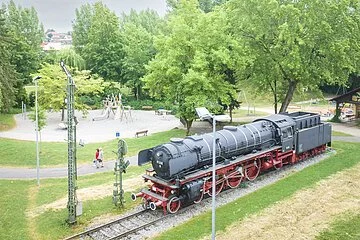 Luftaufnahme der Treuchtlinger Denkmalslok inklusive dahinter liegendem Spielplatz mit Besuchern inmitten von Grün.