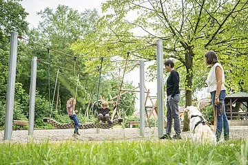 Moderner Spielplatz an der Lok. Zwei kleine Kinder sitzen auf einer geflochtenen Schaukel aus seilen und schwingen. Zwei Mütter stehen mit Hund daneben und beobachten die Kinder. Der Spielplatz befindet sich inmitten einer grünen Kulisse.