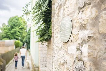 Durch das Bild zieht sich die Marktmauer Treuchtlingens mit angebrachtem Schild. Entlang des daneben verlaufenden Gehsteigs laufen eine Mutter mit Kind im Hintergrund.