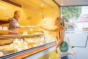 Aufnahme eines Käsestands am Treuchtlinger Wochenmarkt. Eine Frau kauft gerade an der Käsetheke ein. Sie trägt einen Treuchtlinger Stoffbeutel.