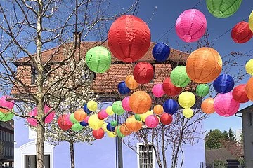 Bunte Lampions über dem Partnerschaftsplatz, im Hintergrund ein blaues Haus und blauer Himmel