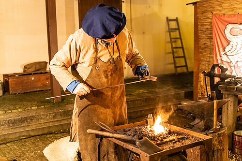 Ein mittelalterlich verkleideter Mann mit Lederschürze steht vor einem Feuer und schmiedet.