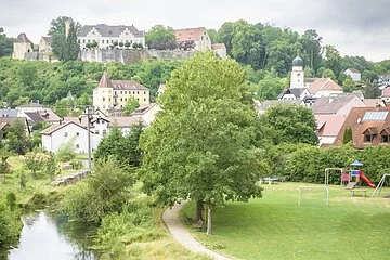 Drohnenaufnahme von Möhren. Im Vordergrund sind 2 Wanderer auf einer Holzbrücke über den Möhrenbach zu sehen. Im Hintergrund thront Schloss Möhren über dem Dorf.