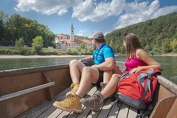 In dem hölzernen Boot sitzen ein Mann und eine Frau, beide in Wanderkleidung mit dem Blick Richtung Kloster Weltenburg gerichtet. Das Kloster ist im Hintergrund von weitem zu sehen umrahmt von grünen Wäldern.