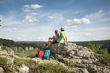 Ein Pärchen sitzt auf einem Felsen. Ihre Wanderrucksäcke lehnen daran. Sie lassen ihren Blick ins Tal schweifen.