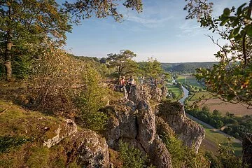 Ein Wanderpärchen, etwas entfernt, steht links auf einem Felsen an der Arnsberger Leite, die gut zu sehen ist. Dahinter erstreckt sich der Blick ins Tal. Der Himmel erstrahlt wolkenlos.