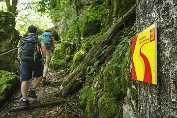 Im Vordergrund hängt ein Wanderschild des Altmühltal-Panoramaweges. Ein Wanderpärchen steigt die Treppen der Riedenburger Klamm hinauf.