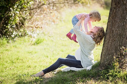 Eine Frau hebt, angelehnt an einen Baum, ein Kind mit beiden Armen in die Luft. Beide haben Spaß und lachen sich an.