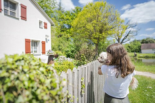 Junges Mädchen , mit einem kleinen weißen Hund auf dem Arm, steht vor einem Holzzaun. Im Hintergrund weißes Haus mit roten Fensterläden.