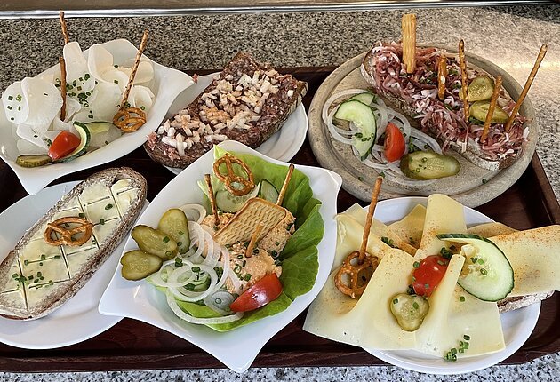 Auf einem Tablett sind verschiedene Brotzeiten wie Butterbrot, Wurstsalat, Käsebrot, Schinkenbrot, Rettich und Tartarbrot zu sehen.
