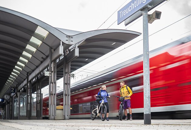 Aufnahme an einem Treuchtlinger Bahnsteig. Im Hintergrund fährt ein Regionalzug der DB durch den Bahnhof. Am Gleis stehen zwei Radfahrer mit ihren E-Bikes.