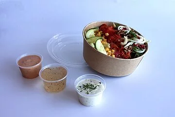Ein gemischter Salat mit Gurke, Mais, Tomate, Blattsala, Nüssen und Granatapfelkernen. Der Salat befindet sich in einem Behältnis für Außerhausverkauf. Es gibt verschiedene Dressings zur Auswahl.