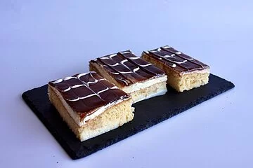 Drei Stück Kuchen vom Blech mit Creme und Schokoguss angerichtet auf schwarzer Schiefertafel.