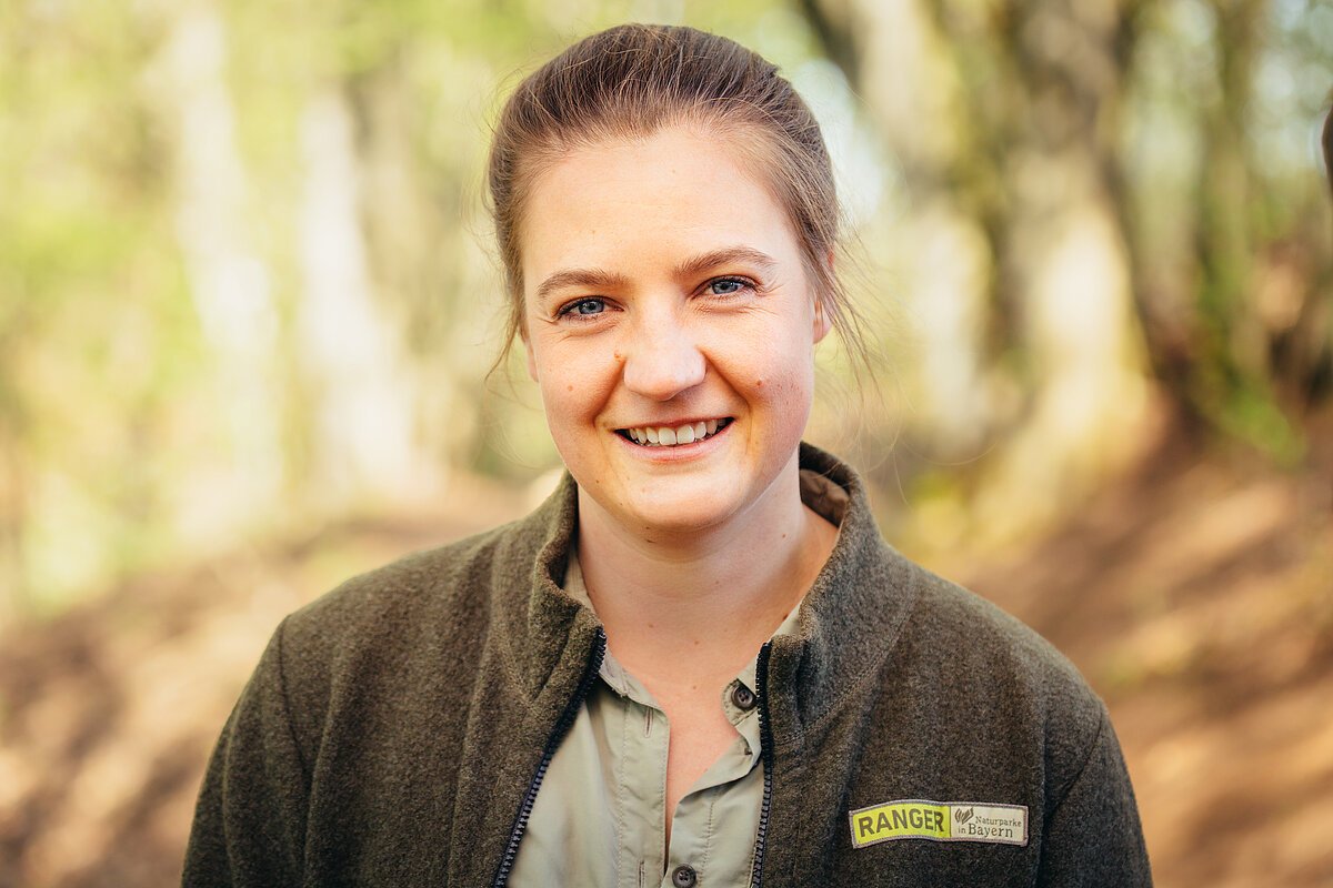 Eine junge, braunhaarige Frau trägt eine waldgrüne Ranger-Jacke und schaut in die Kamera. Im Hintergrund der Wald.