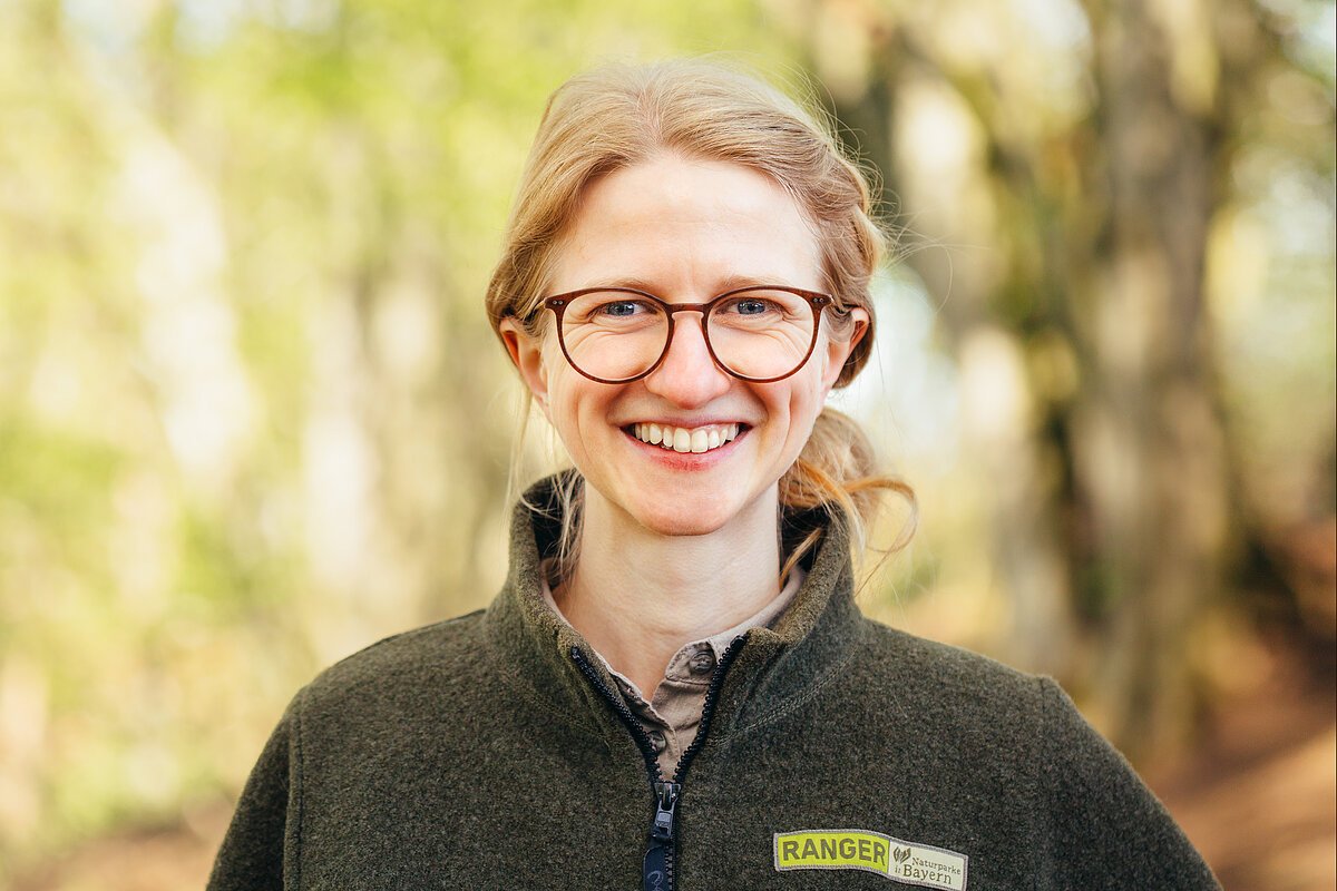 Eine junge, blonde Frau mit Brille und geflochtenem Haar trägt eine waldgrüne Ranger-Jacke und schaut in die Kamera