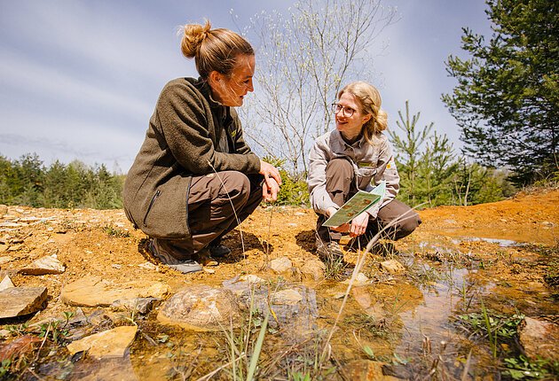 Zwei Naturpark-Rangerinnen in Outdoorkleidung knien vor einer Pfütze im steinigen Untergrund