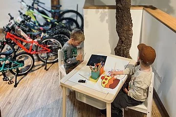 Ein Kindertisch, an dem zwei Jungen sitzen. Links stehen verschieden farbige Kinderfahrräder