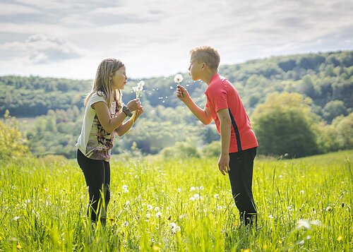 Zwei Kinder auf einer Wiese mit Pusteblumen in den Händen, die sie gemeinsam pusten