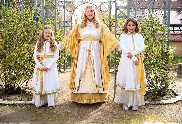 Das Christkind in einem Engelskleid und die Engel in weniger üppigen Kleidern, stehen nebeneinander vor einem Gartenatelier, mit Sonnenschein im Hintergrund. Das Christkind hat ihre Arme ausgebreitet hinter den Engeln.