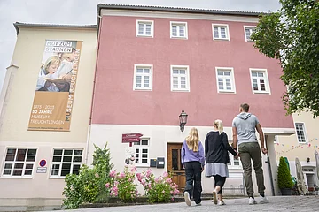 Frontansicht des Museums Gebäudes inklusive Banner des 50-Jahre Jubiläums. Im Vordergrund läuft eine Familie in Richtung Museumseingang.