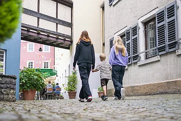 3 Kinder laufen aus Richtung des Blauen Hauses zum Museumseingang. Die historische Häuserverbindung und das pinke Museumsgebäude sind im Hintergrund zu sehen.