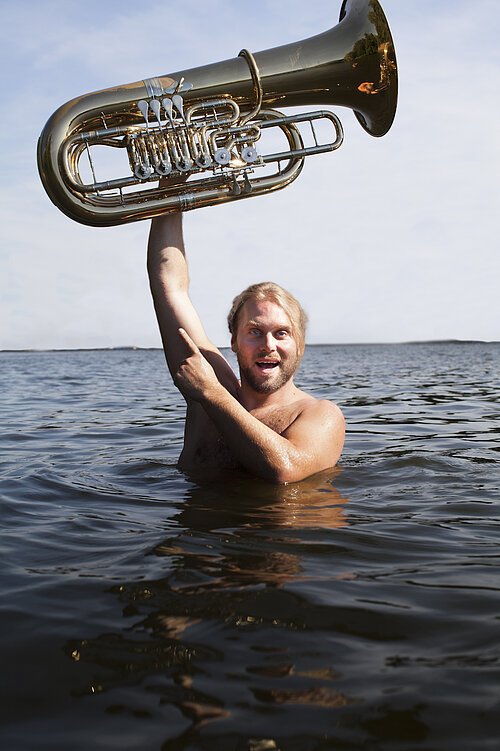 Auf dem Bild ist Andreas Martin Hofmeir in einem See zu sehen, wie er seine Tuba nach oben streckt und mit seiner anderen Hand darauf zeigt.