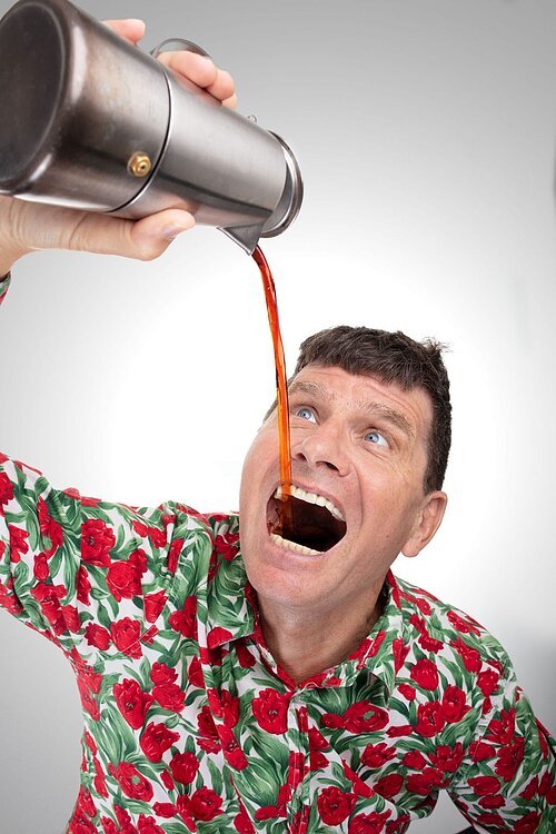 Auf dem Bild ist der Kabarettist Mäc Härder zu sehen, wie er eine rot braune Flüßigkeit, möglicherweise Bier, aus einer silbernen Kaffeekanne trinkt.