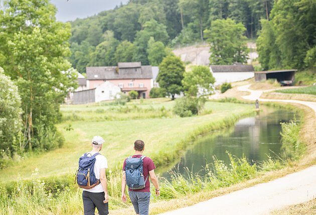 Zwei Wanderer laufen entlang eines Schotterweges durch das Möhrenbachtal. Im Hintergrund sind Mühlen und ein bewaldeter Berg zu sehen.