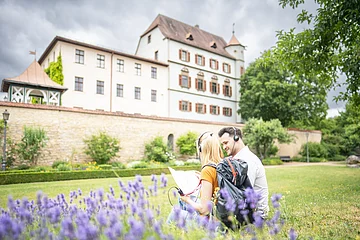 Auf der Wiese vor dem Stadtschloss sitzen 2 Touristen. Sie lauschen der Treuchtlinger Stadttour über die Kopfhörer der Audioguides.