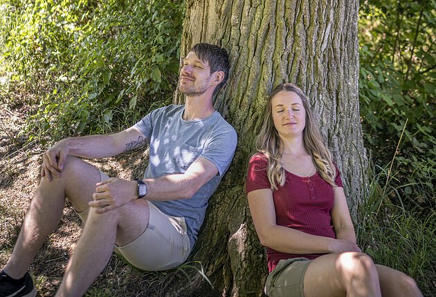 Ein Mann und eine Frau sitzen am Waldboden und lehnen an einem Baumstamm